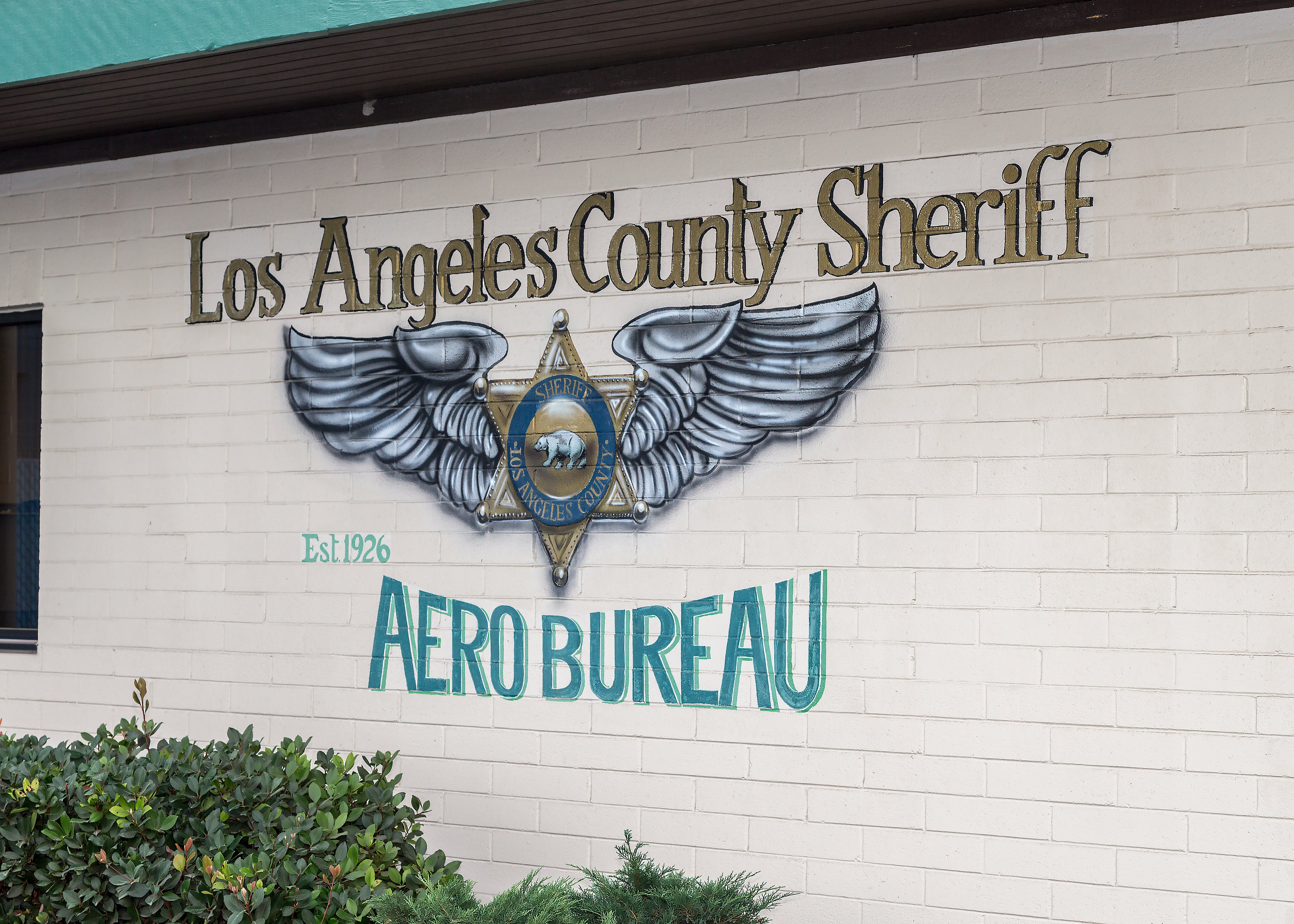 Los Angeles County Sheriff Aero Bureau painted sign on a white brick building wall, featuring a winged badge emblem and teal lettering.