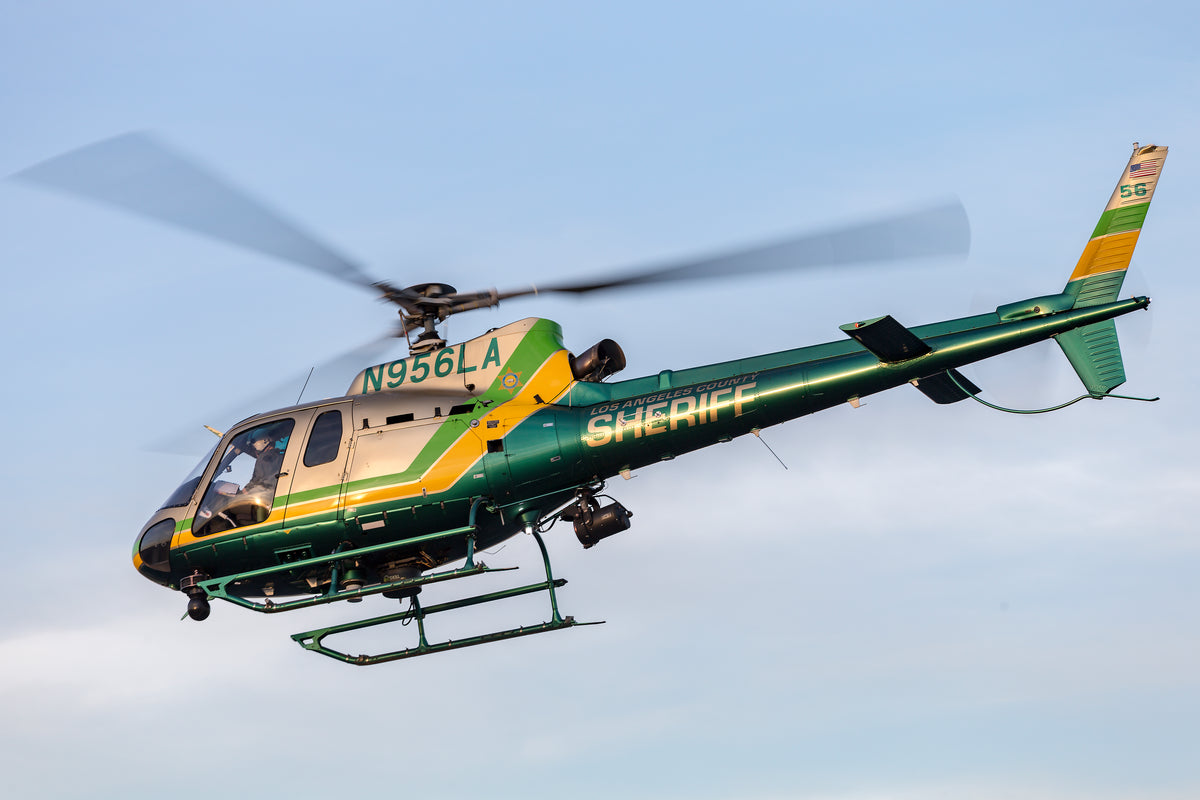 A green and tan helicopter with 'SHERIFF' and registration 'N956LA' printed on it, captured in flight against a pale blue sky.