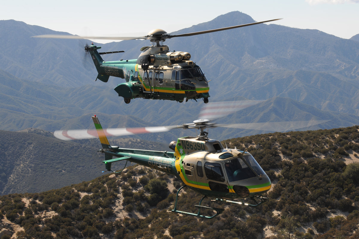 Two helicopters flying over a mountainous desert landscape. The helicopters are olive green, white, and yellow, with blurred rotor blades visible due to motion.